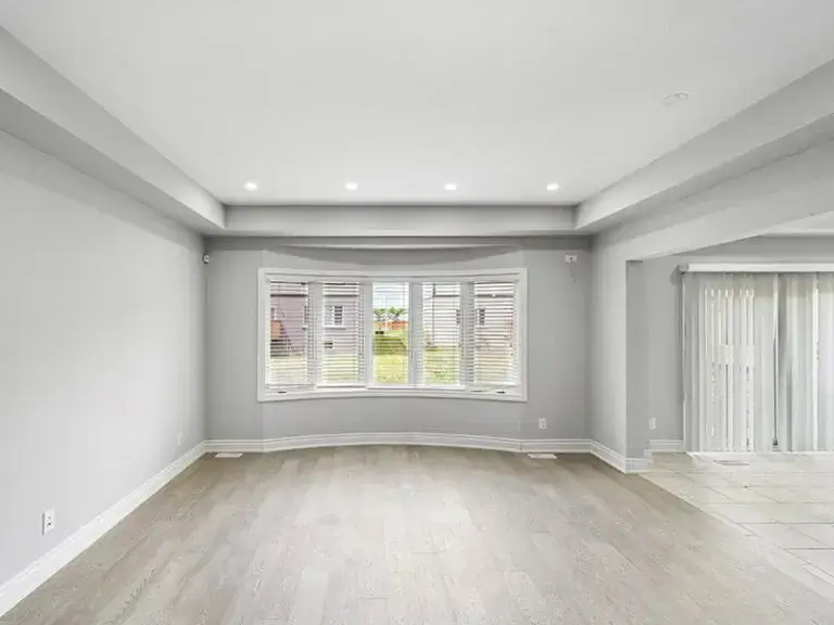 Empty living room with light hardwood floors, gray walls, bay window with blinds, recessed lighting, and tray ceiling